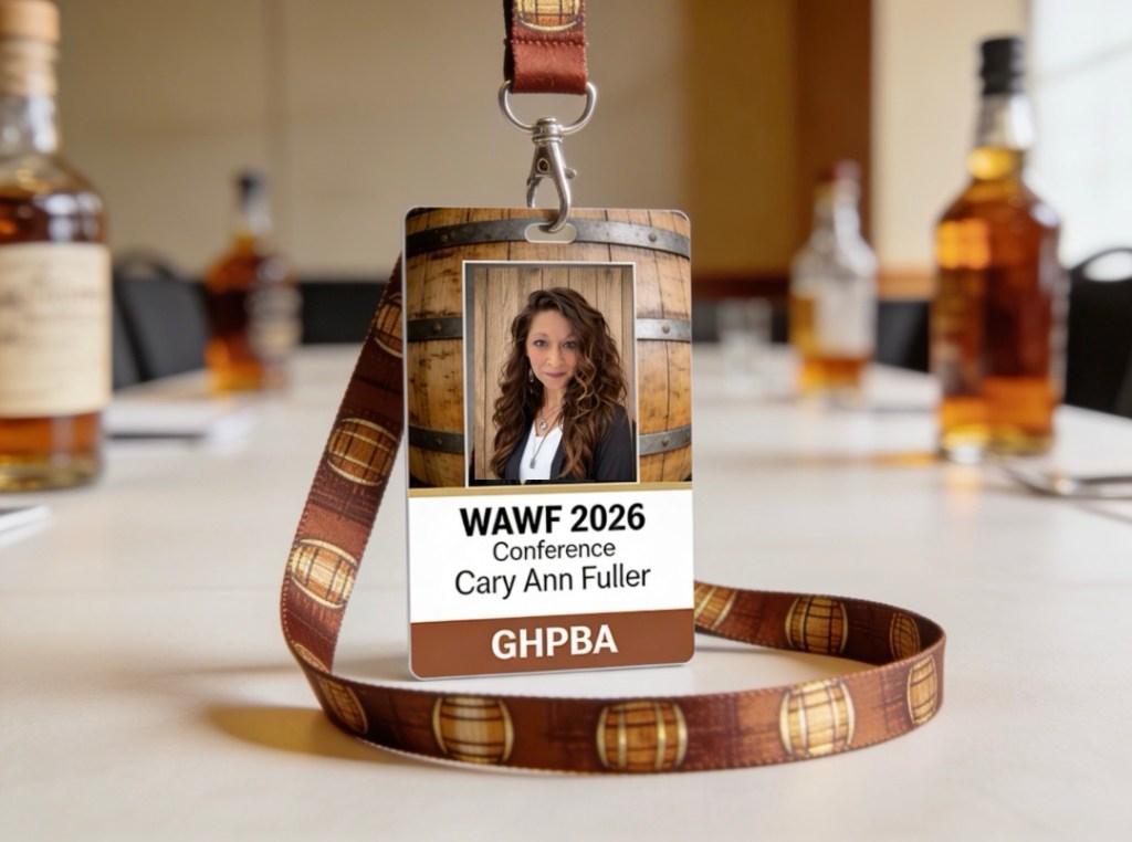 A conference name tag for WAFF 2026 featuring a photo of a woman named Cary Ann Fuller, displayed on a lanyard with a barrel design. In the background, there are bottles of whiskey on a table.